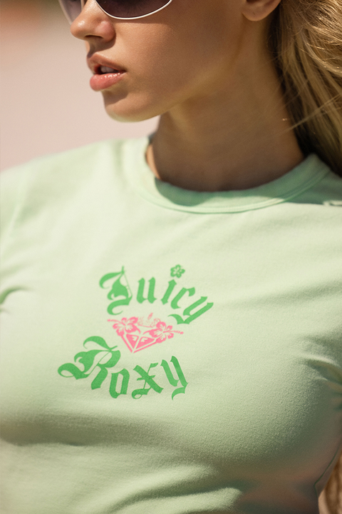 A person in sunglasses wears a Roxy x Juicy Baby Tee, a light green fitted short sleeve shirt with a pink floral graphic on the front. The cropped image shows only the lower face and torso.