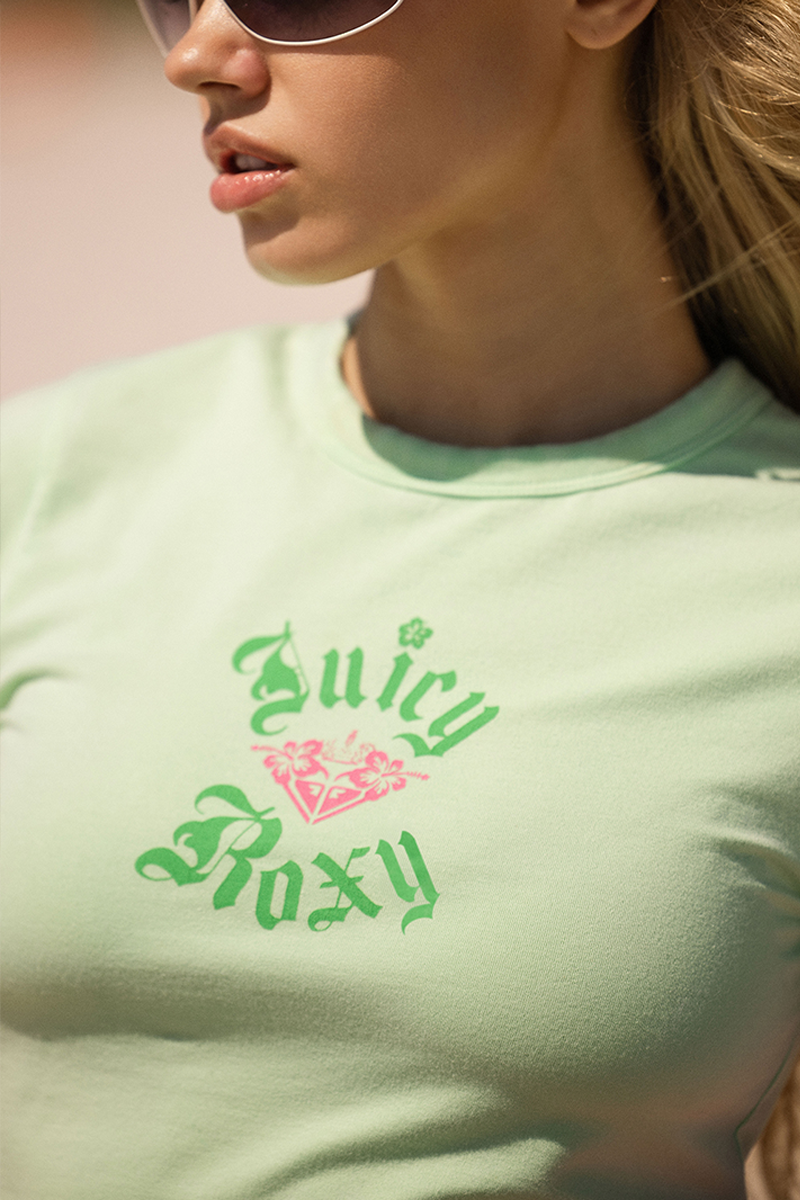 A person in sunglasses wears a Roxy x Juicy Baby Tee, a light green fitted short sleeve shirt with a pink floral graphic on the front. The cropped image shows only the lower face and torso.