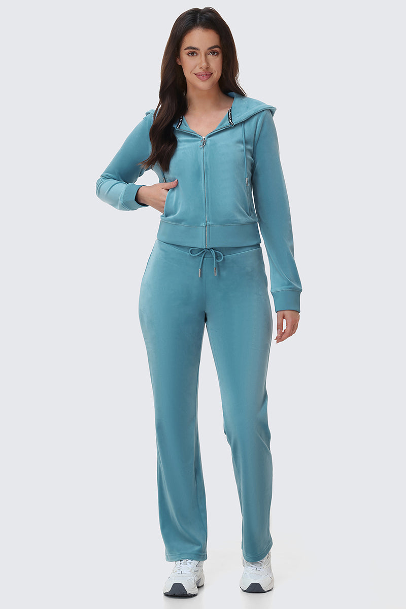 A woman wearing a matching teal Y2K bedazzled OG Big Bling Velour Hoodie with thumbholes and straight-leg sweatpants, paired with white athletic shoes.