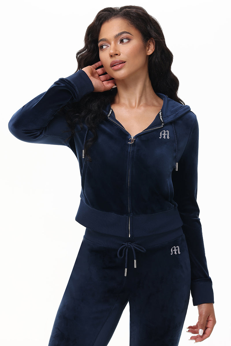 A woman with long wavy hair poses in a Custom Initial OG Big Bling Velour Hoodie and matching joggers, featuring classic Y2K style and rhinestone-inspired details. She stands sideways with one hand near her neck, evoking vintage Juicy vibes.
