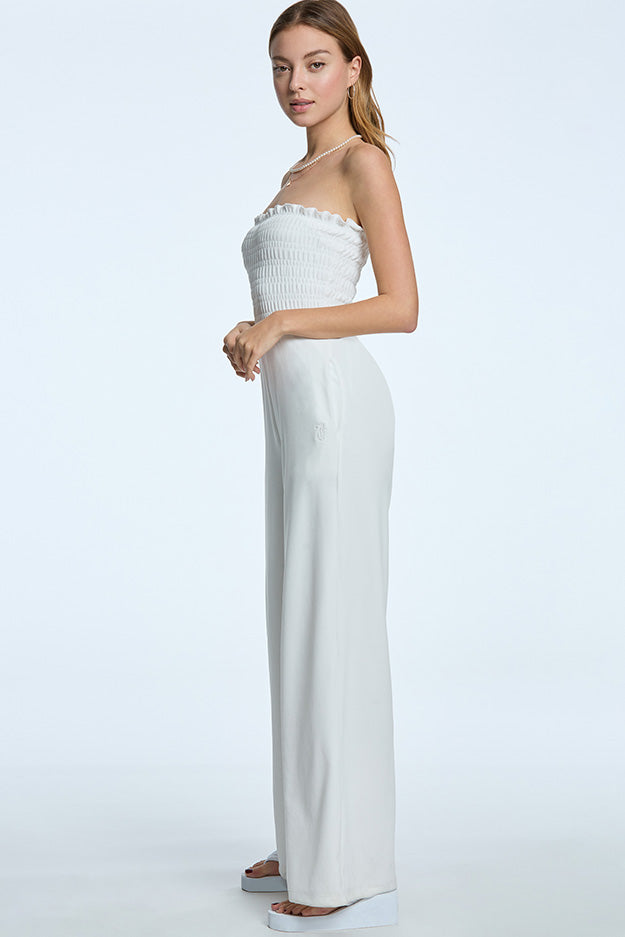 A young woman with straight, light brown hair stands sideways against a plain background, wearing sandals and the white Smocked Velour Jumpsuit featuring a pull-on style with wide-leg pants.