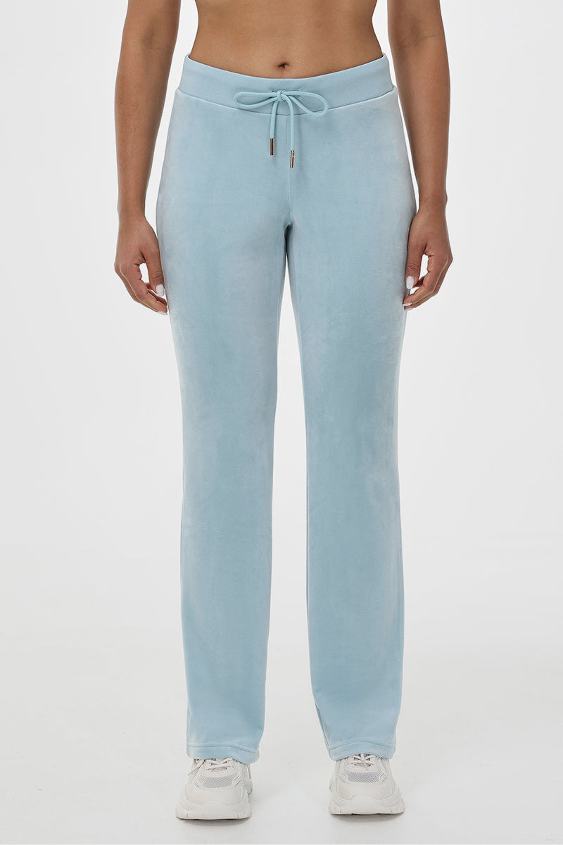 A person shown from shoulders to feet wears the OG Big Bling Velour Track Pants in light blue, featuring a drawstring waist, paired with white sneakers and standing against a plain white background.