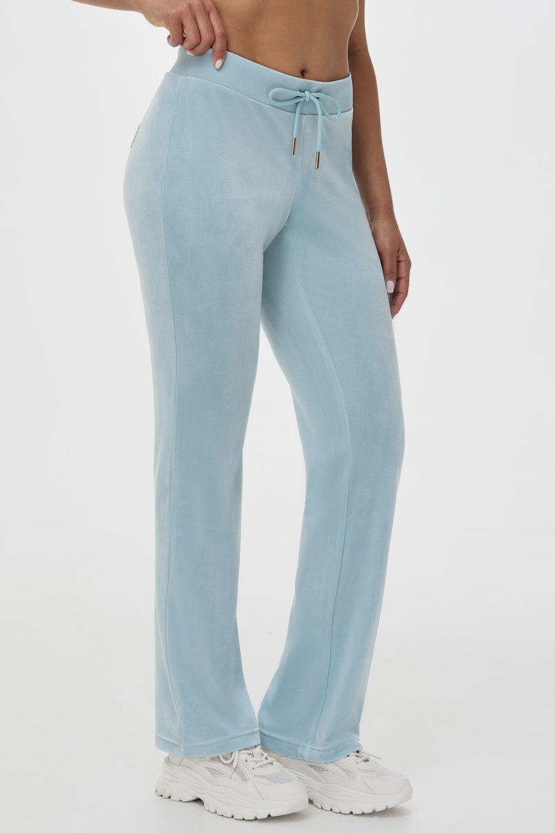A person wearing OG Big Bling Velour Track Pants in light blue with a subtle Juicy embellishment and white sneakers, standing against a plain white background.