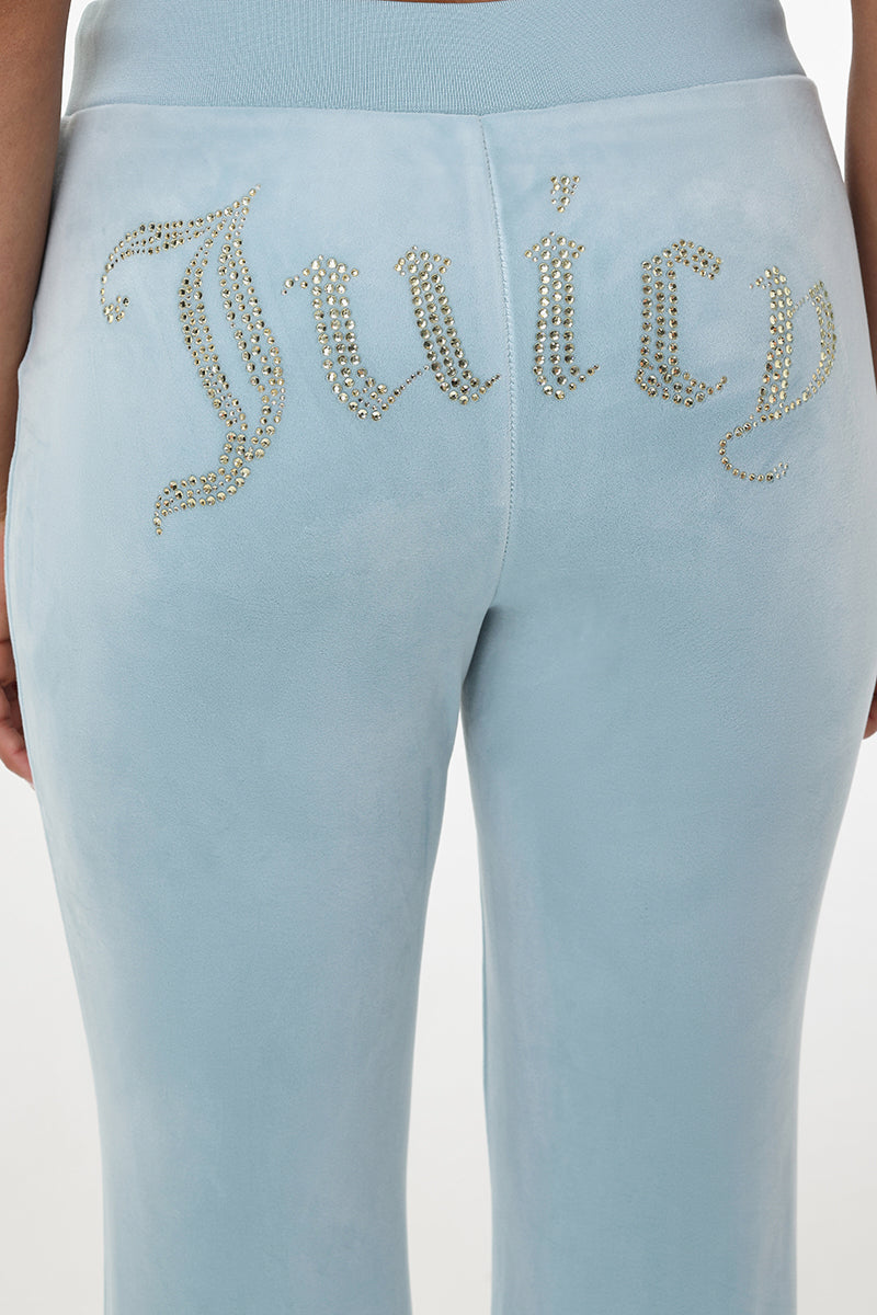 A person wearing the OG Big Bling Velour Track Pants in light blue, channeling a Y2K vibe with a sparkling “Juicy” embellishment spelled out in rhinestones across the back.