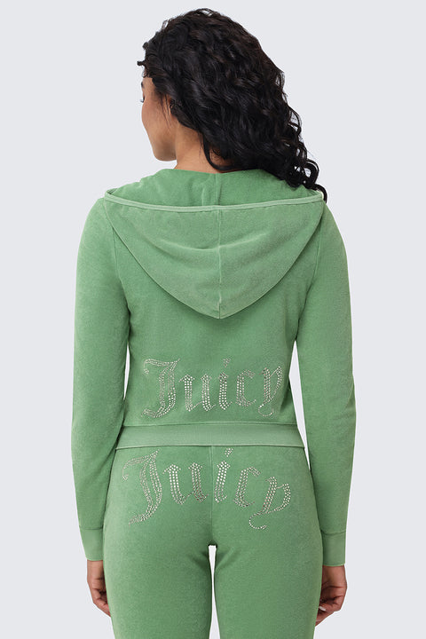 A woman with long, wavy hair is seen from the back wearing the Big Bling Washed Terry Hoodie in green, featuring a sparkling Juicy logo on the back of the jacket and across the seat of her matching pants.