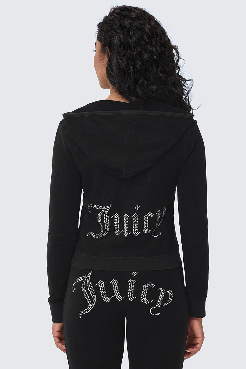 A woman with curly hair faces away, wearing the Big Bling Washed Terry Hoodie and black pants. Both feature the word "Juicy" in sparkling rhinestone logo letters across the back.