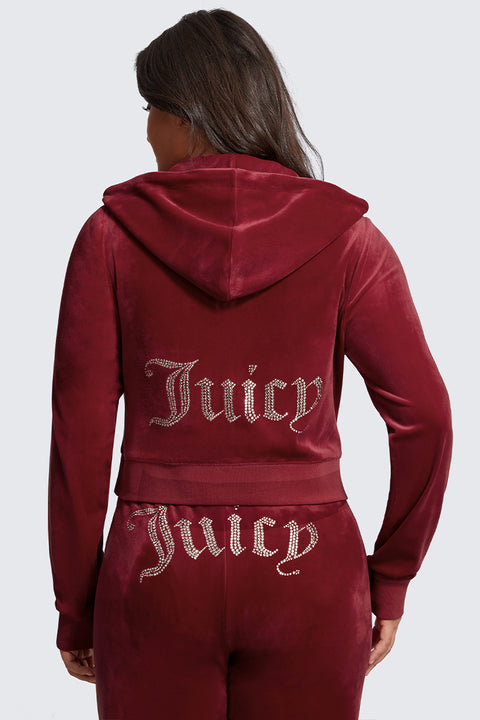 A woman in a plus-size OG Big Bling Velour Hoodie, maroon with thumbholes and rhinestone Juicy lettering on the back, stands facing away from the camera.