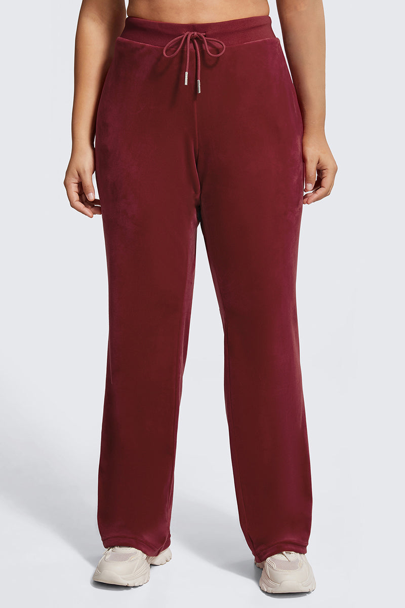 A person models the Plus-Size OG Big Bling Velour Track Pants in burgundy with a drawstring and light sneakers, standing against a plain light gray background. The cropped image showcases a subtle Y2K vibe.
