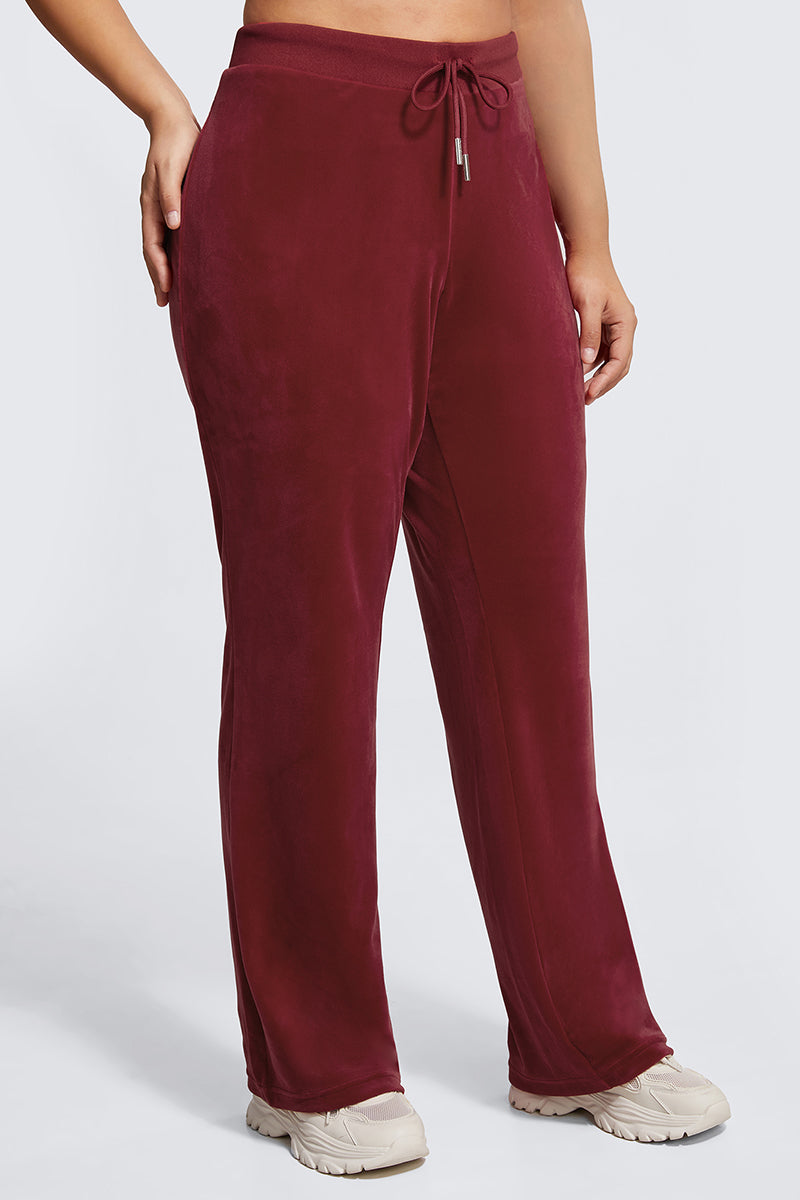 A person wears Plus-Size OG Big Bling Velour Track Pants in burgundy with a drawstring waist and cream sneakers, standing against a plain light gray background. The photo is cropped from shoulders to feet.