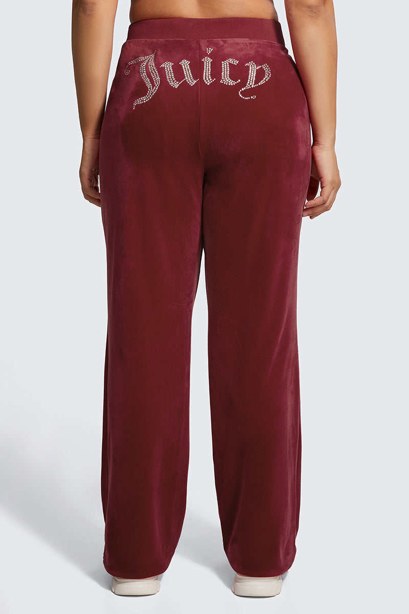 A person seen from the back wears the Plus-Size OG Big Bling Velour Track Pants in burgundy, featuring sparkling rhinestone Juicy embellishment on the waistband, paired with white sneakers.