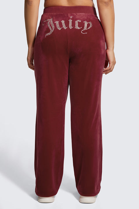 A person seen from the back wears the Plus-Size OG Big Bling Velour Track Pants in burgundy, featuring sparkling rhinestone Juicy embellishment on the waistband, paired with white sneakers.