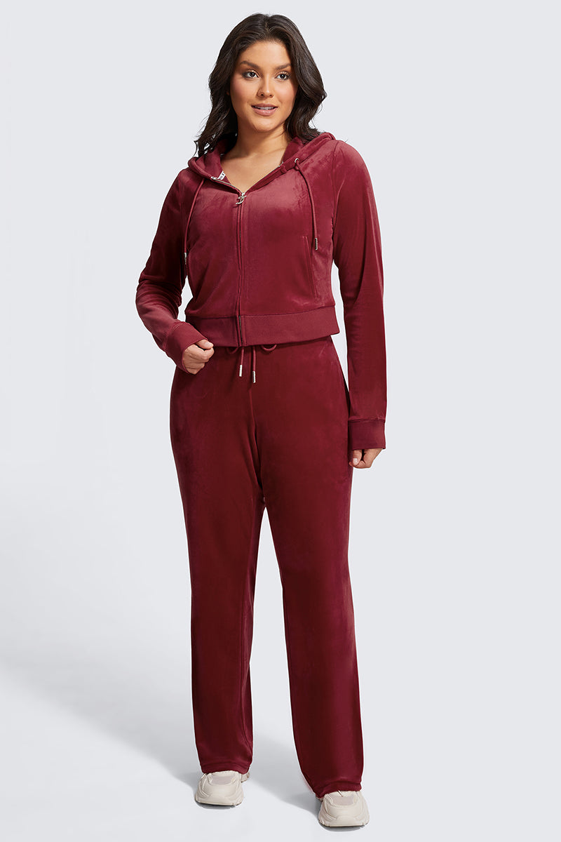 A woman with long brown hair and white sneakers smiles confidently in Plus-Size OG Big Bling Velour Track Pants and a matching burgundy zip-up hoodie, posing against a plain light gray background.