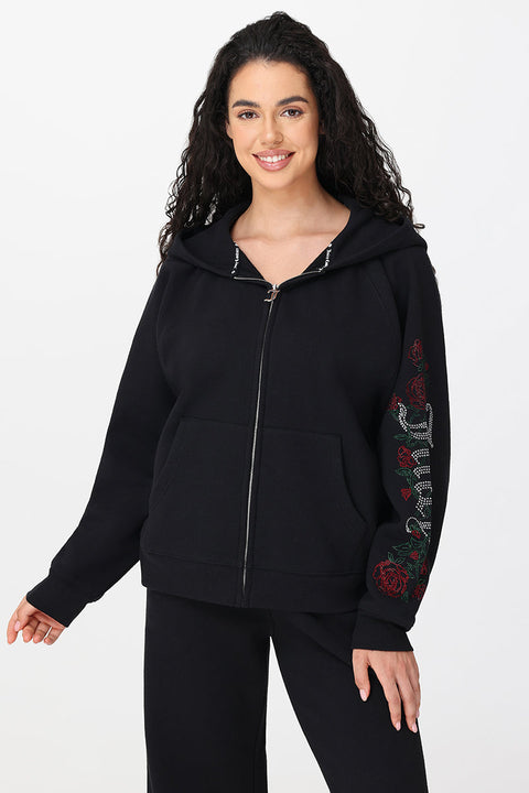 A woman with long curly hair smiles against a white background, wearing the Roses Oversized Zip Front Hoodie featuring red rose embroidery, sparkling text on one sleeve, and matching black pants.