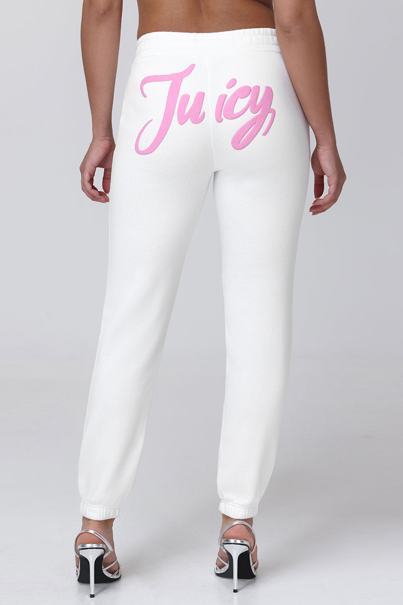 A person stands facing away, wearing Sweetheart Fleece Sweatpants in a relaxed fit with "Juicy" in pink script across the back, paired with white high-heeled sandals and a bare back.