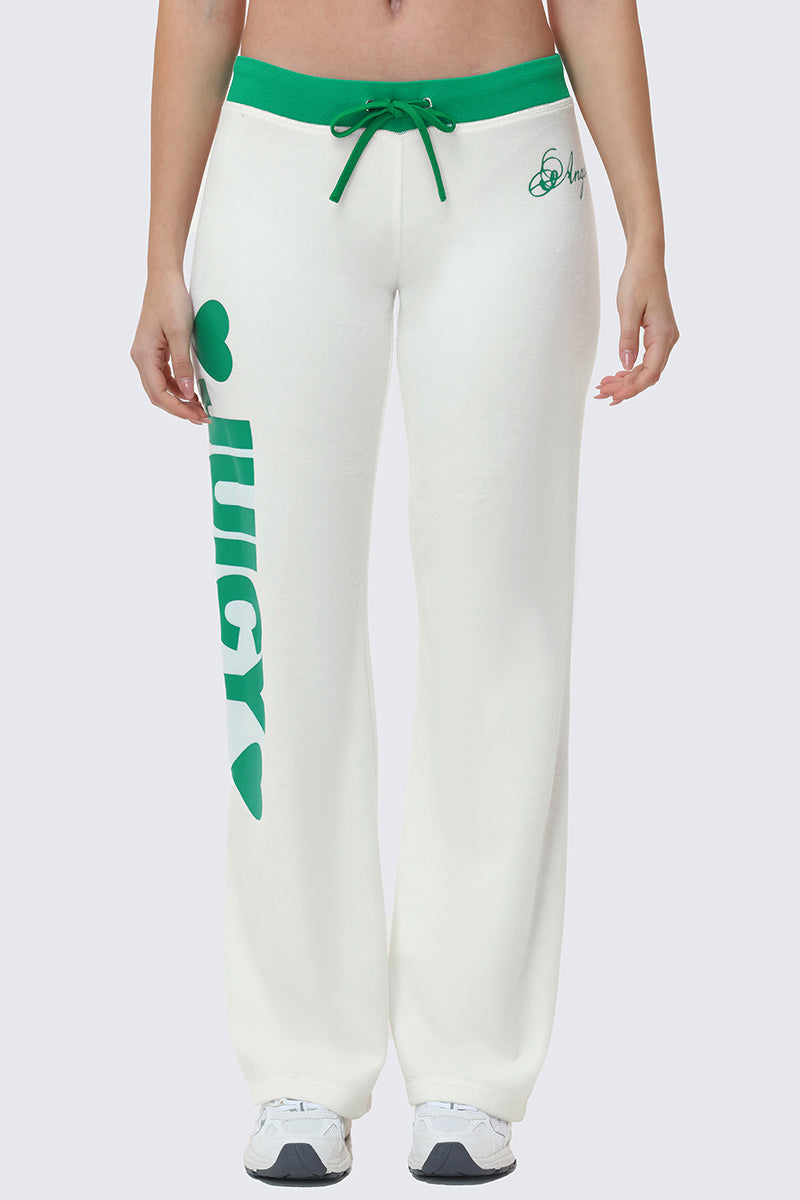 A person wearing Contrast Low Rise Track Pants in white with green trim, relaxed fit, green drawstring, and “JUICY” in bold green letters with a heart printed vertically on one leg; paired with white sneakers.
