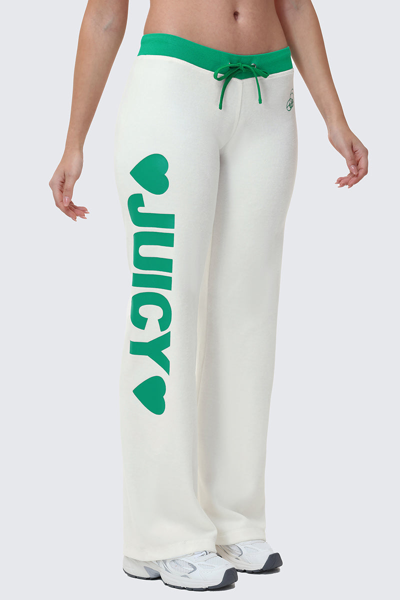 A person wears Juicy’s Contrast Low Rise Track Pants—white with green trim, a green drawstring, and bold JUICY text with hearts on one leg—paired with white sneakers for a sporty loungewear look.
