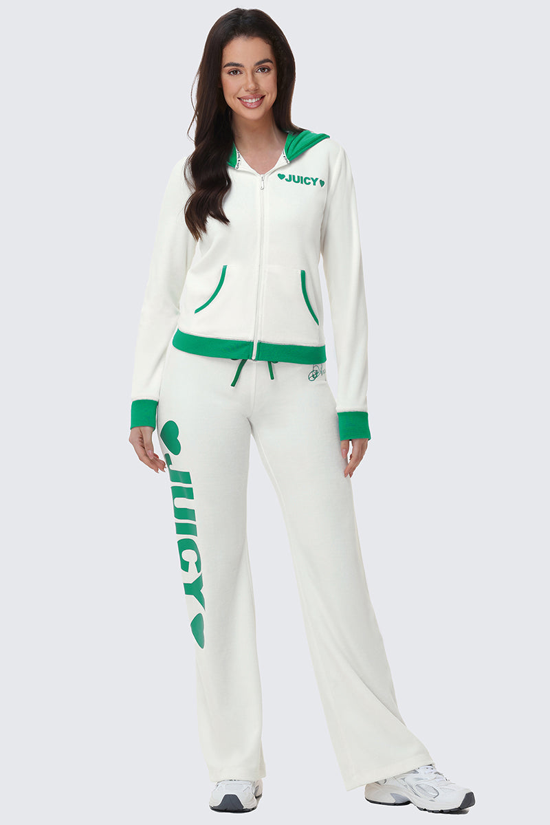 A woman smiles in sporty loungewear: white Contrast Low Rise Track Pants with green trim and a bold leg logo, paired with a matching relaxed-fit jacket featuring “JUICY” on the chest. She completes the look with white sneakers.