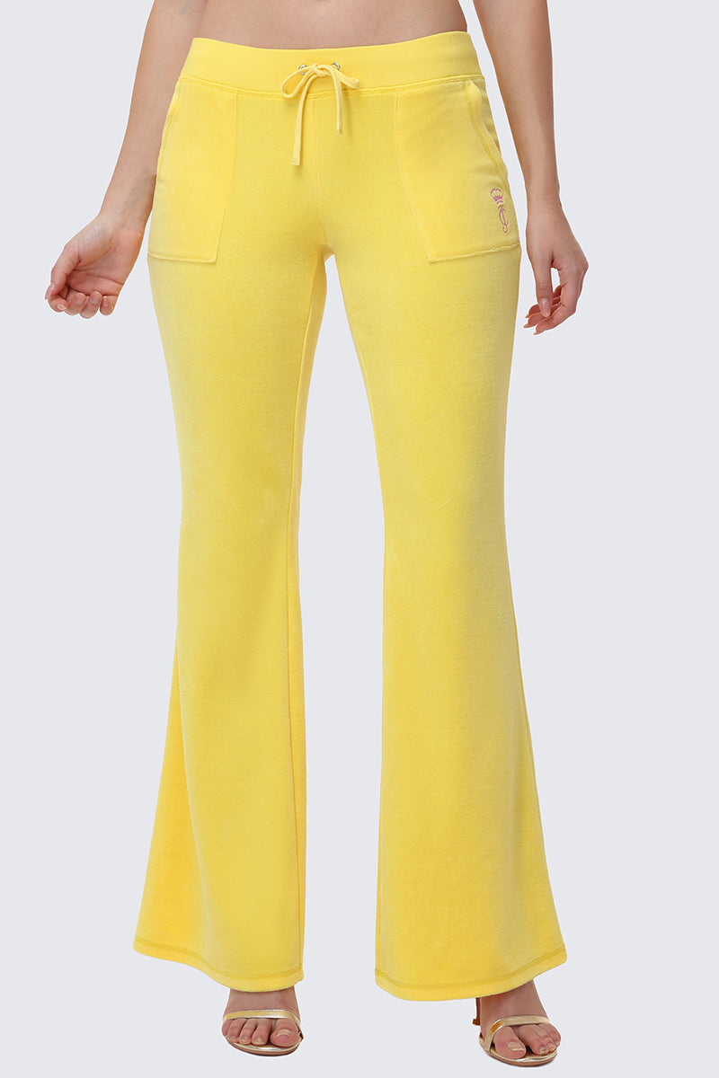 A person wears the Heritage Cotton Velour Track Pants in yellow, featuring front patch pockets and a drawstring waist, standing against a plain light gray background with heeled sandals. Only the lower body is shown; classic Juicy vibe.