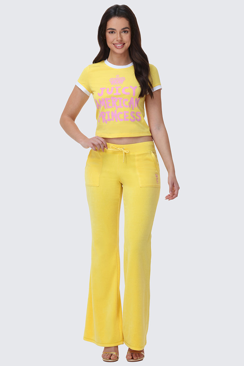A woman smiles in yellow Heritage Cotton Velour Track Pants with front pockets and a drawstring waist, paired with a matching crop top reading “Juicy American Princess” in pink letters.