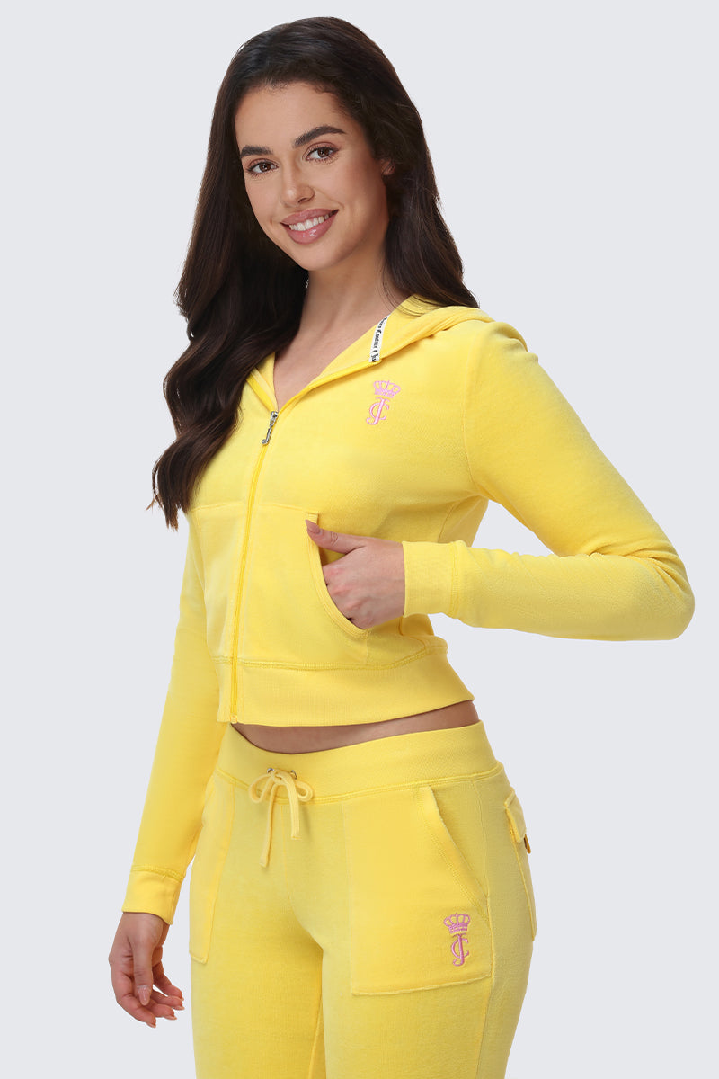 A woman with long brown hair smiles in a bright yellow American Princess Heritage Hoodie and matching drawstring pants, both featuring a small pink crown logo, standing against a light gray background.