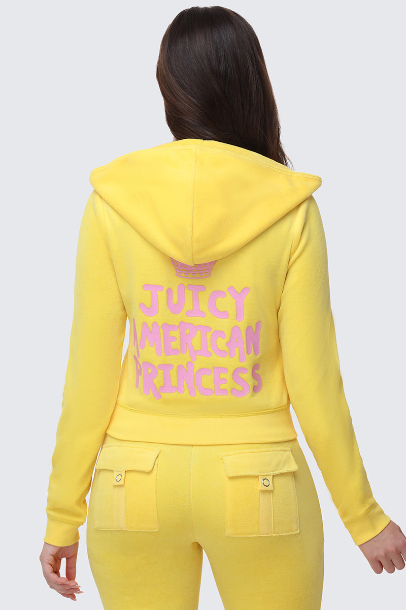 A woman facing away wears the American Princess Heritage Hoodie, a yellow velour zip hoodie and tracksuit. "JUICY AMERICAN PRINCESS" in pink with a crown logo decorates the back, and the pants feature two flap pockets with buttons.