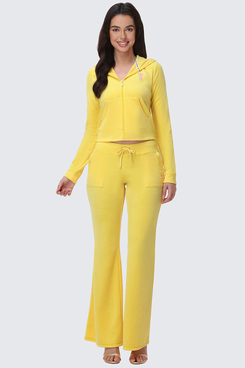 A woman with long dark hair smiles while wearing the American Princess Heritage Hoodie in yellow velour and matching wide-leg drawstring pants, both featuring the Juicy crown logo. She pairs the outfit with open-toed heels against a plain light background.