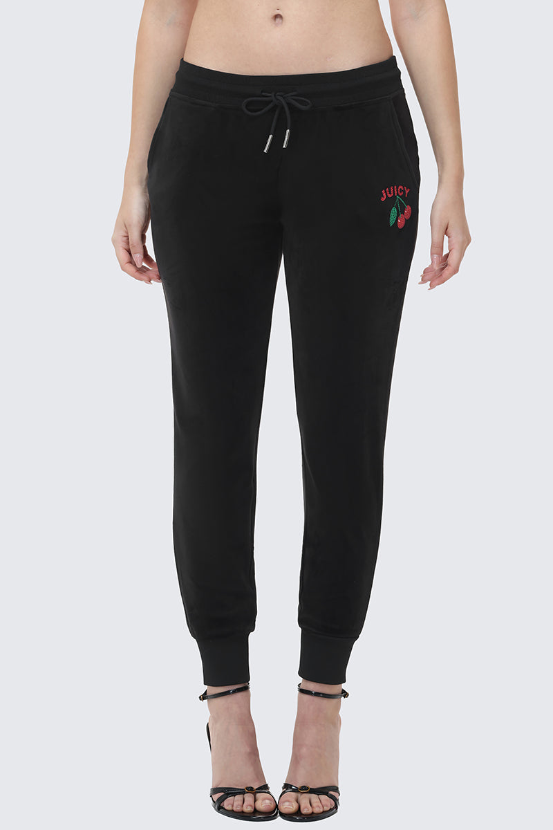 A woman wears the Cherry Bling Velour Jogger featuring a drawstring waist, cuffed ankles, and a Juicy cherry graphic on the left hip, paired with black heeled sandals. Her midriff is visible.