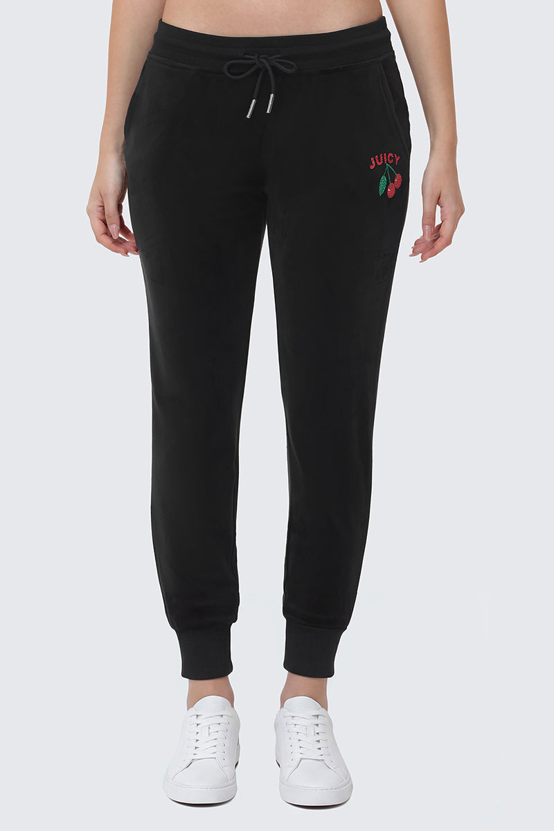 A person wears Cherry Bling Velour Jogger—black velour pants with a drawstring waist, small JUICY logo, and cherry detail on the left thigh—paired with white sneakers. Image shows from shoulders down.