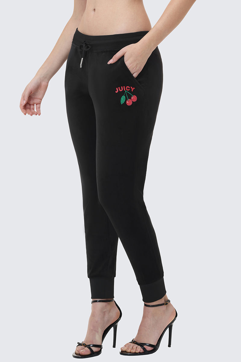 Woman in Cherry Bling Velour Jogger with Juicy graphic and cherry embroidery on the left pocket, featuring a drawstring waist and ankle cuffs, styled with black high-heeled sandals for a trendy, comfortable look.
