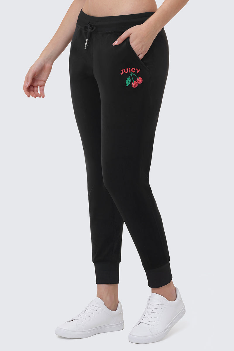 Someone wears the Cherry Bling Velour Jogger with a small JUICY graphic and cherries on the pocket, plus a drawstring waist, styled with white sneakers, standing against a plain light background.