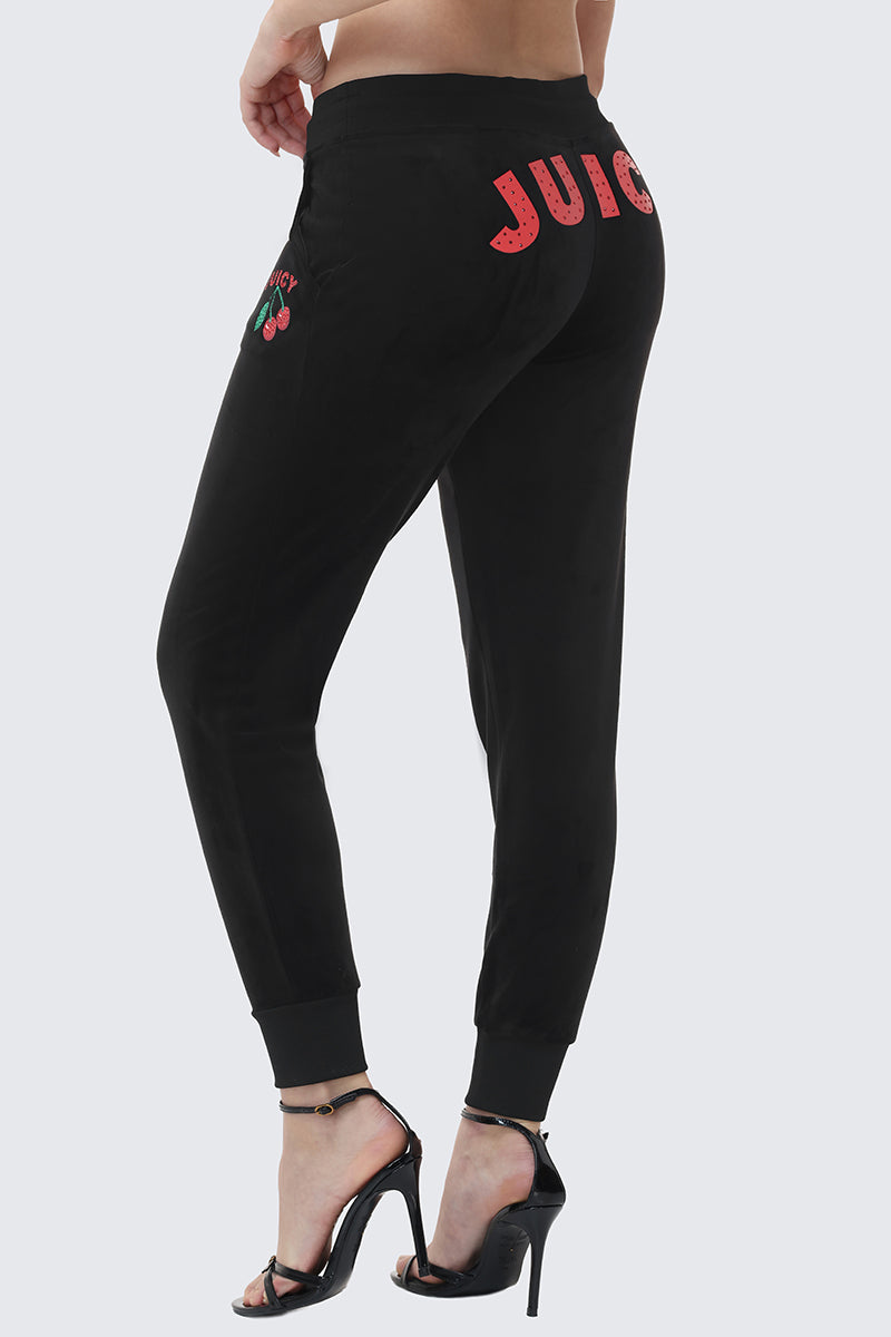 A person wearing the Cherry Bling Velour Jogger, featuring a drawstring waist and bold JUICY graphic with two cherries on the back, paired with black high-heeled sandals against a light gray background.