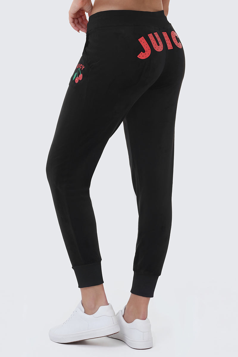 A person is shown from the back wearing the Cherry Bling Velour Jogger—black joggers with "JUICY" in red letters on the back, a cherry Juicy graphic on the side, a drawstring waist, and paired with white sneakers.