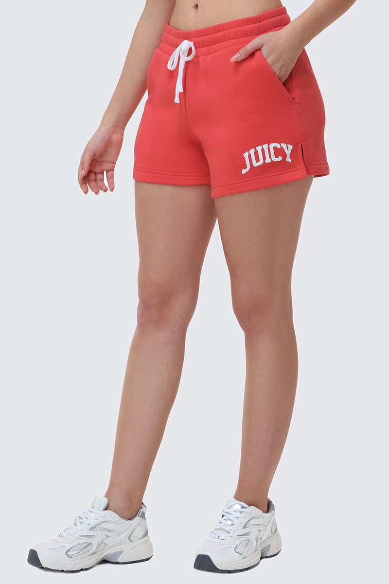Someone is shown from the waist down wearing Collegiate Booty Shorts in red with a Juicy logo on the left leg, paired with white sneakers and a white drawstring, standing against a plain light background.