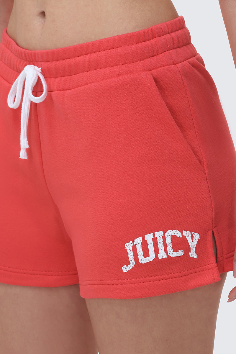 A person wearing Collegiate Booty Shorts in bright red terry cloth, featuring a white drawstring and bold white Juicy logo on the left leg. The photo is cropped to show only the shorts and part of the torso.