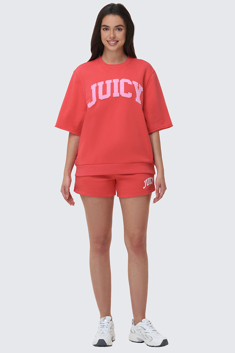 A woman faces forward, smiling in Collegiate Booty Shorts with "JUICY" on the left leg, paired with a matching red short-sleeve sweatshirt and white sneakers.