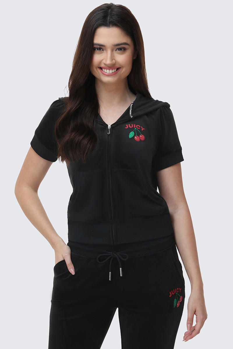 A woman with long dark hair smiles while wearing the Cherry Bling Velour Hoodie, featuring a Juicy cherry graphic and "JUICY" embroidery. She has one hand in her pocket.