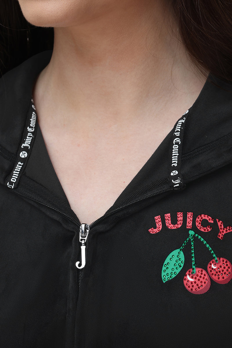 Close-up of a woman wearing the Cherry Bling Velour Hoodie, a black short-sleeve hoodie with a Juicy cherry graphic adorned with sequins and rhinestones on the chest. The hood drawstrings display "Juicy Couture" printed in white.