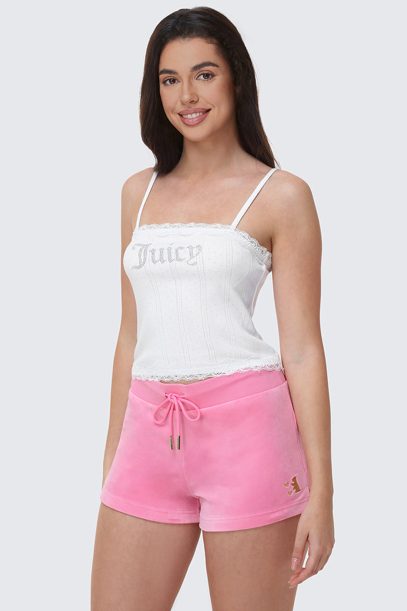 A woman smiles while wearing the Bling Lace Trim Cami in white with pink drawstring shorts featuring gold lettering on the left side, standing against a plain light gray background.