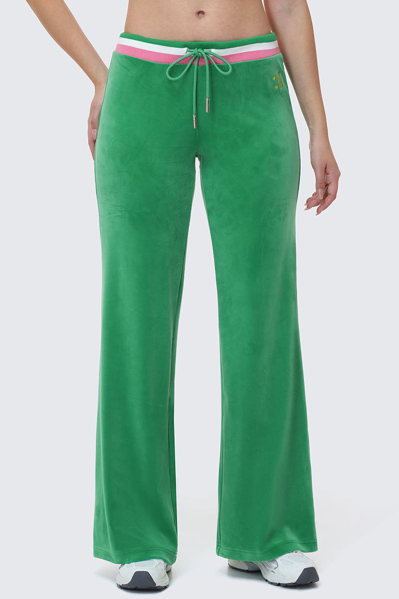 A person wears the Heritage Stripe Bling Velour Track Pants featuring green velour fabric, a pink and white striped waistband with drawstring, paired with white sneakers. Only the lower half of their body is visible.