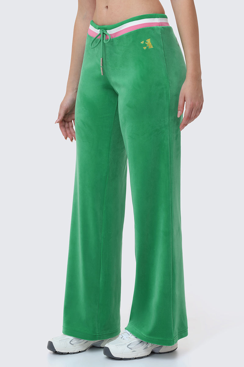 Someone stands against a plain light background wearing Heritage Stripe Bling Velour Track Pants in bright green with a striped waistband, small logo on the left hip, and white athletic shoes.