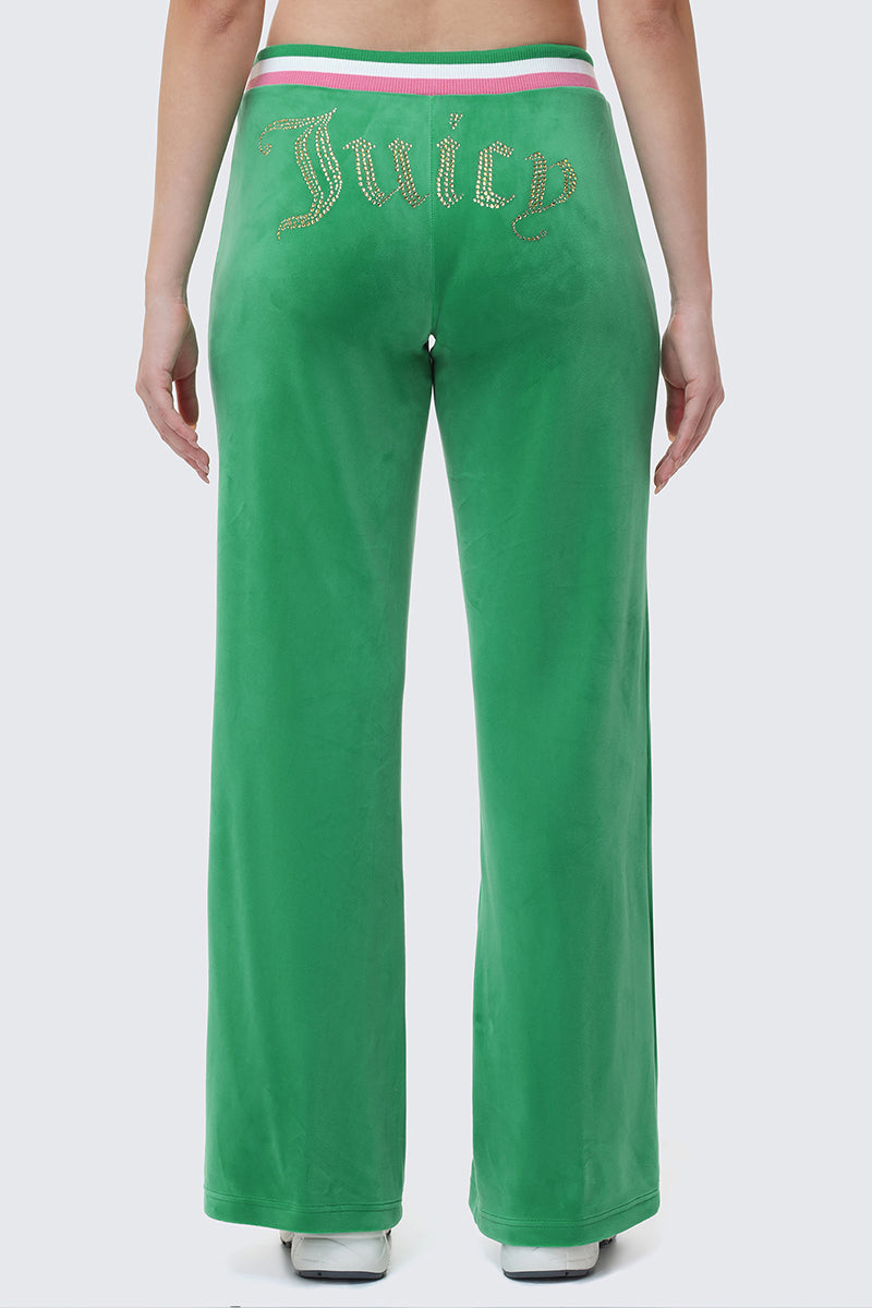 A person models the Heritage Stripe Bling Velour Track Pants, featuring a rhinestone Juicy logo across the back and a striped waistband with pink, white, and green accents, standing with their back to the camera.