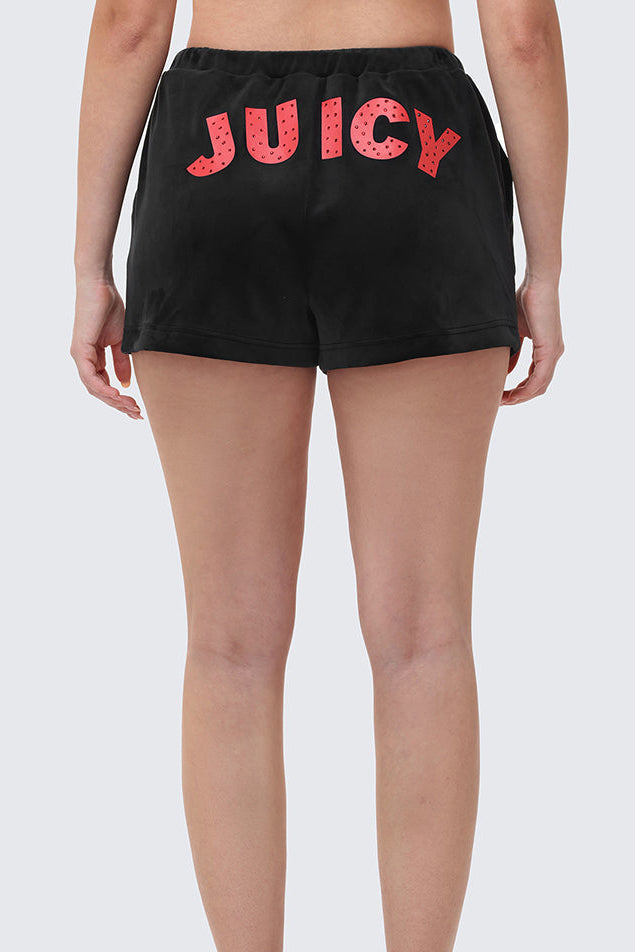 A person wearing Cherry Bling Velour Shorts featuring a pink Juicy graphic on the back, standing with their back to the camera and paired with black high-heeled sandals.