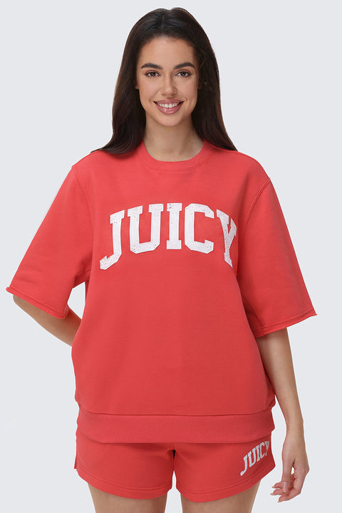 A woman with long dark hair smiles in a red Collegiate Sweatshirt and matching shorts with “JUICY” on the hem, perfect for everyday wear, standing against a plain light background.