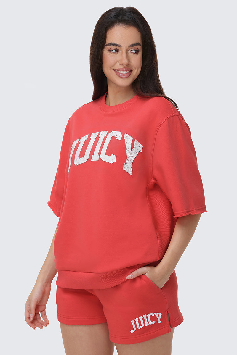 A woman smiles in a red short-sleeve Collegiate Sweatshirt and matching shorts set, both featuring bold white "JUICY" lettering—perfect for standout everyday style. She stands with one hand in her pocket.