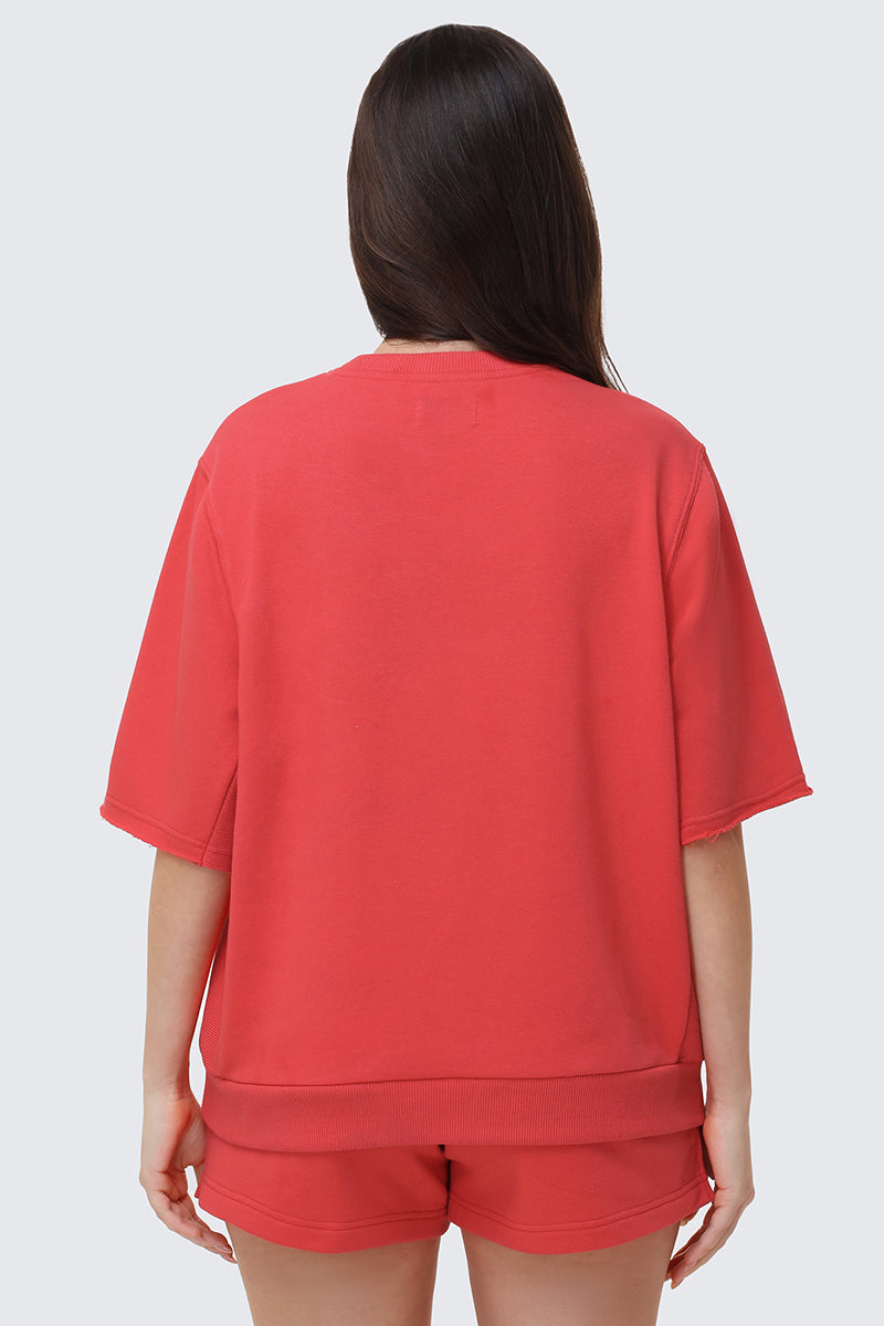 A person with long brown hair is shown from the back, wearing a loose-fitting red Collegiate Sweatshirt and matching red shorts, standing against a plain light gray background.