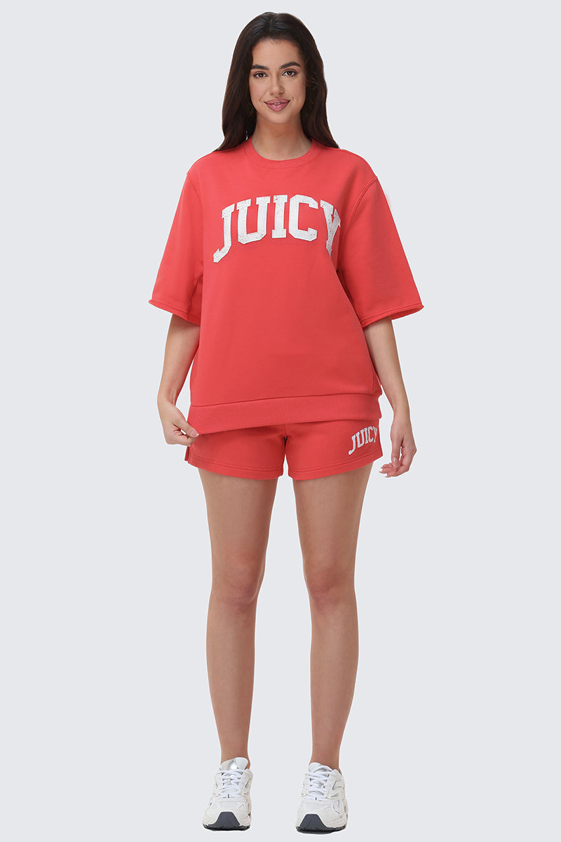 Against a light gray background, a woman wears a red Collegiate Sweatshirt with bold white lettering and matching shorts, completing the everyday look with white sneakers and a bright smile.