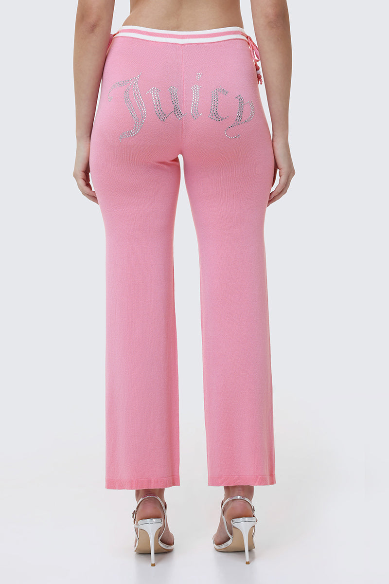 A person wearing Big Bling Wide Leg Pants in pink velour with a rhinestone logo across the back, paired with white high-heeled sandals, is shown from behind against a light background.