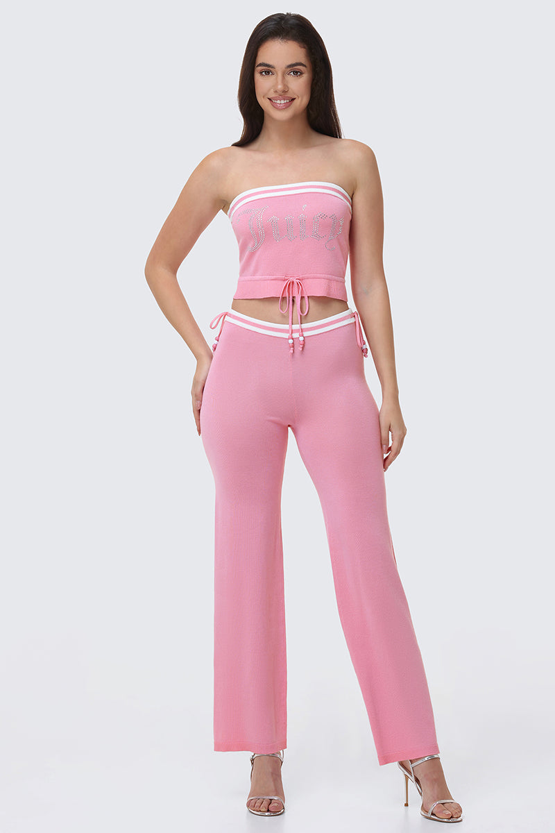 A woman smiles in Big Bling Wide Leg Pants featuring a pink flare style and contrast striped waistband, paired with a pink rhinestone Juicy logo tube top and silver heels. She has long dark hair.