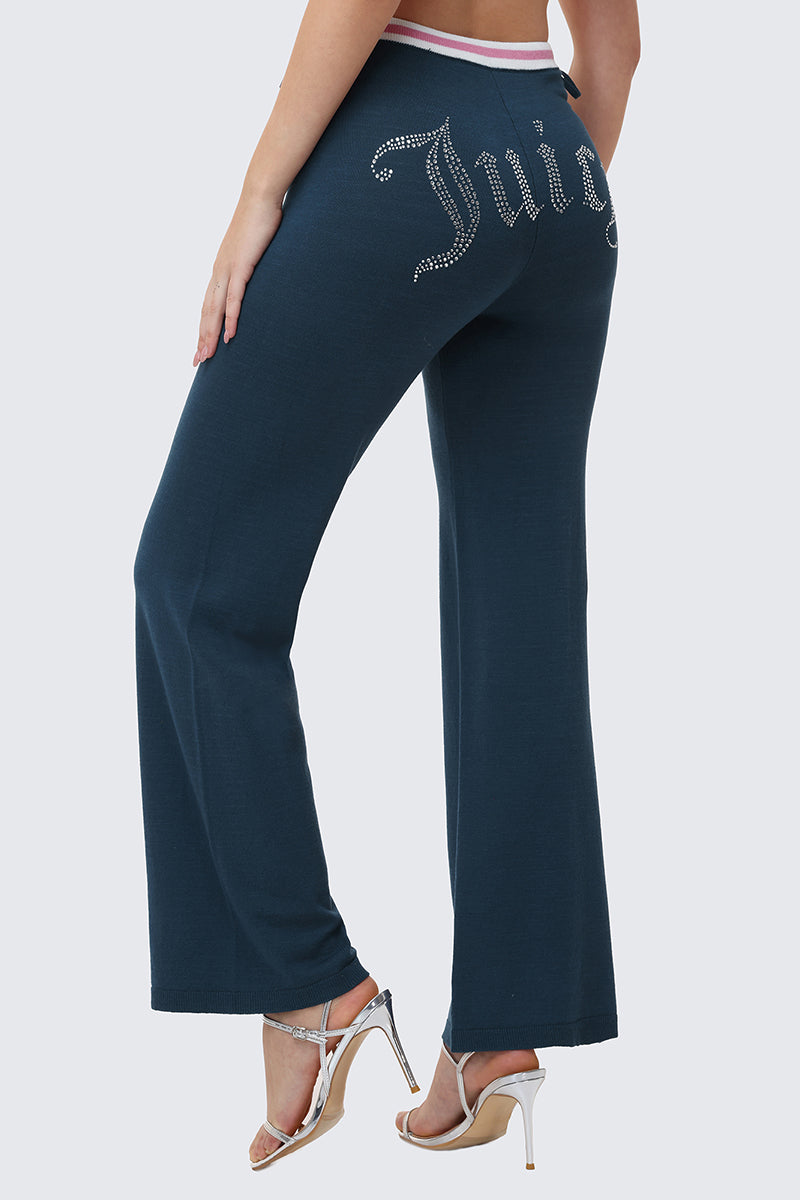 A woman wears Big Bling Wide Leg Pants in dark blue velour, featuring a rhinestone Juicy logo on the back and a pink-and-white contrast striped waistband, styled with high-heeled sandals.