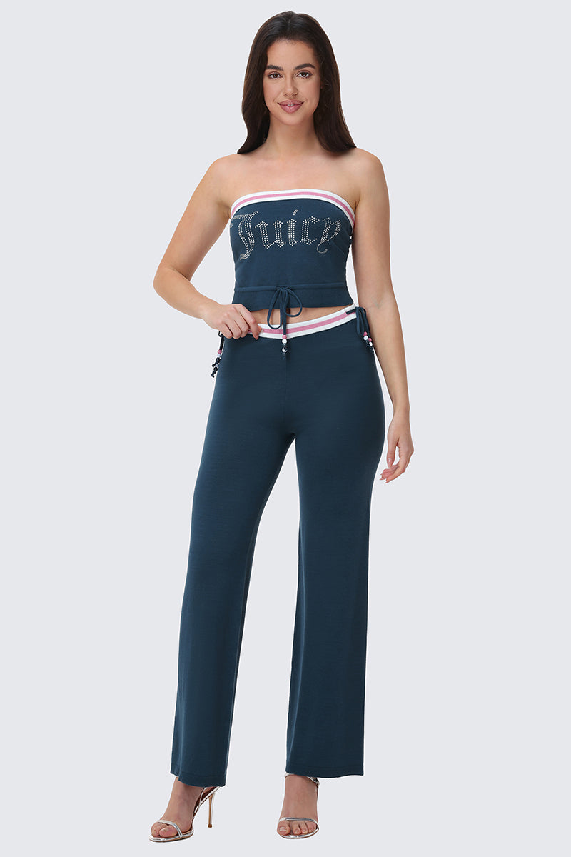 A woman smiles at the camera in heels, wearing Big Bling Wide Leg Pants—a strapless, dark teal two-piece with the Juicy logo on top, contrast striped waistband, white and pink trim, drawstring waist, and ribbed wide-leg pants.